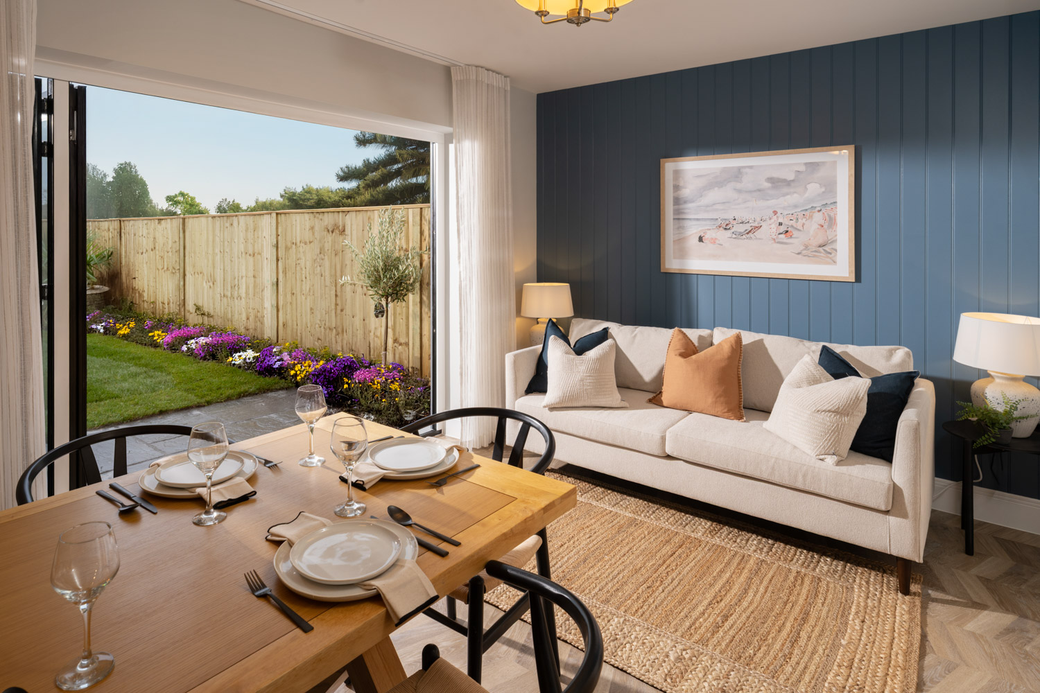 Open plan dining area with dining table and chairs at the middle with dark blue far wall and cream sofa to the rear. Patio doors open to the rear garden